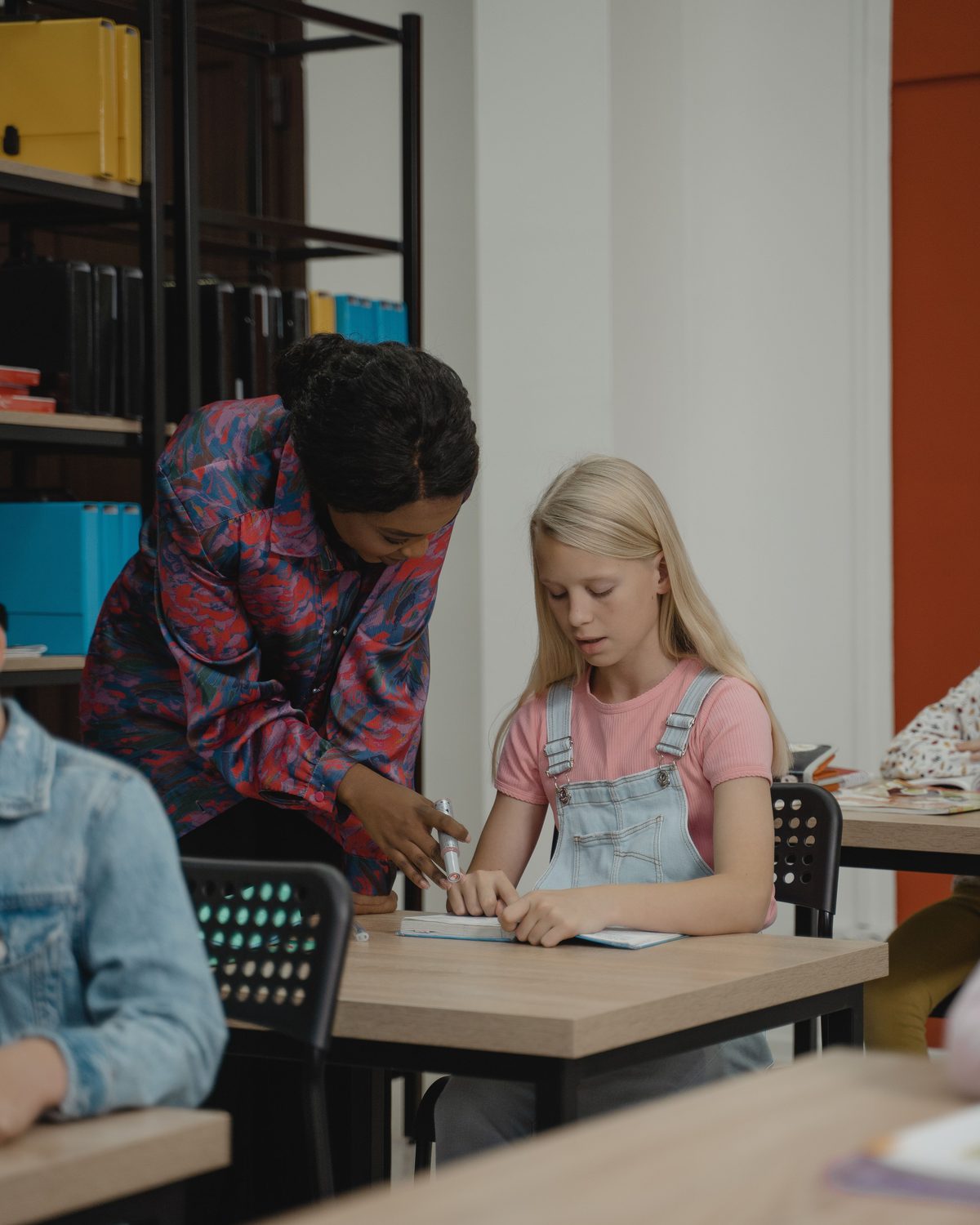 Eine Schulbegleiterin unterstützt eine Schülerin am Tisch, im Hintergrund weitere Kinder im Klassenraum.